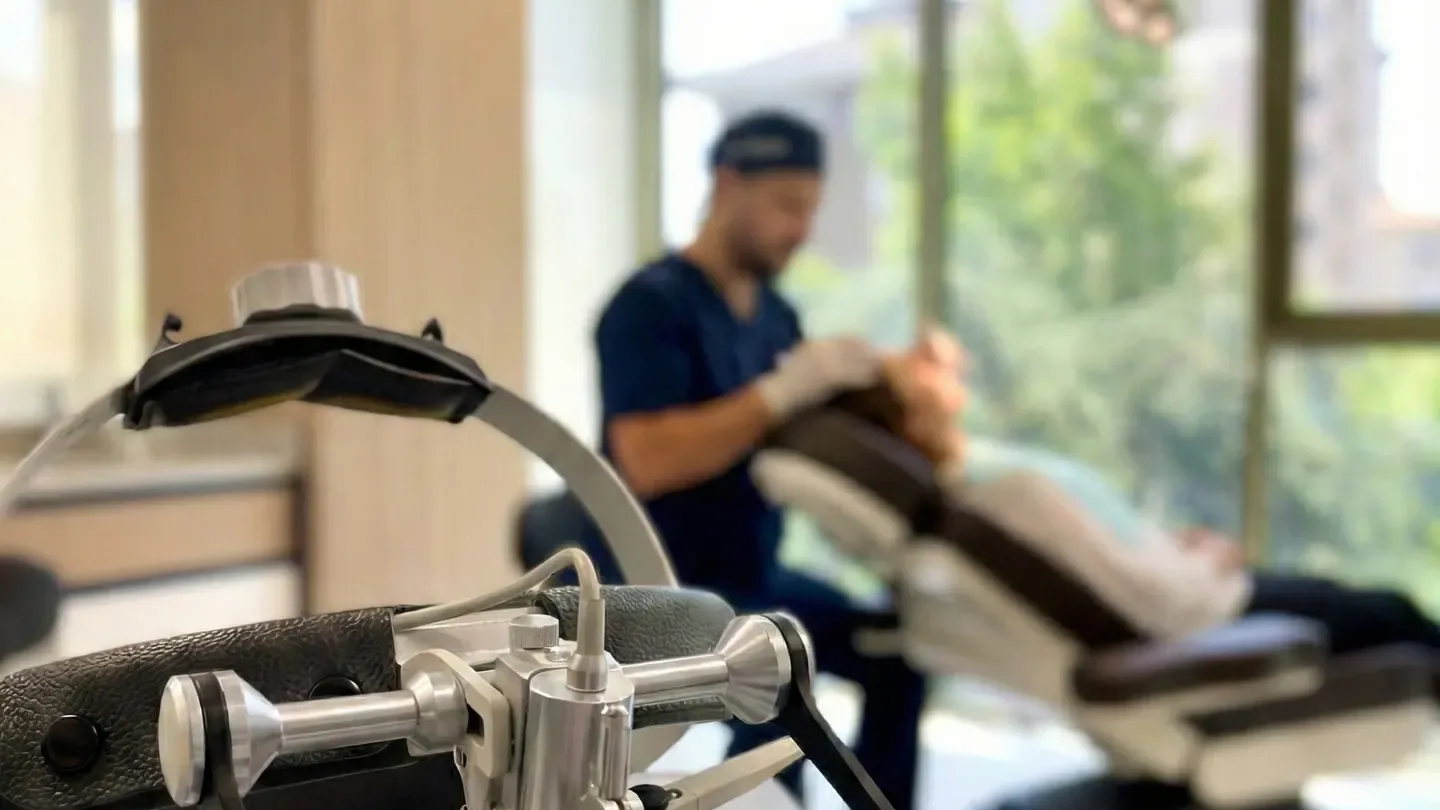 Dr. Mesut Demir working on a single scheduled hair transplant in the Pure Line operating room in Istanbul, with surgical loupes resting in the foreground and daylight coming through the full-height windows behind the treatment chair